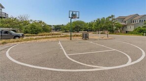 Una cancha de baloncesto al aire libre con un aro, rodeada de árboles y edificios residenciales.