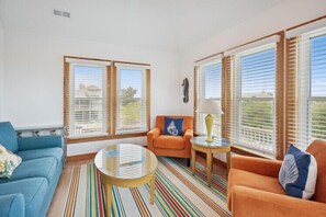 Sala de estar con dos sofás, una mesa de centro y una lámpara. Amplias ventanas con persianas.