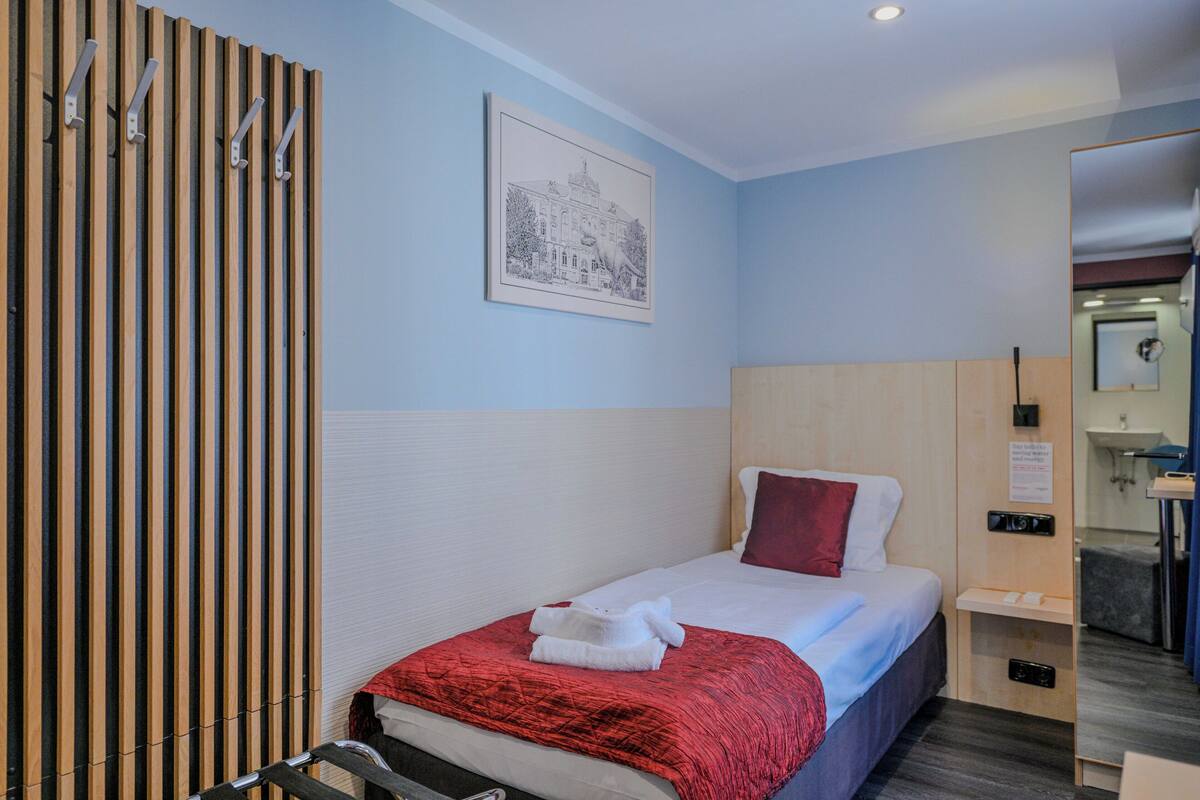 A hotel room with a bed, a red blanket, a white pillow, a wooden panel wall, a framed picture, and a small bedside table.