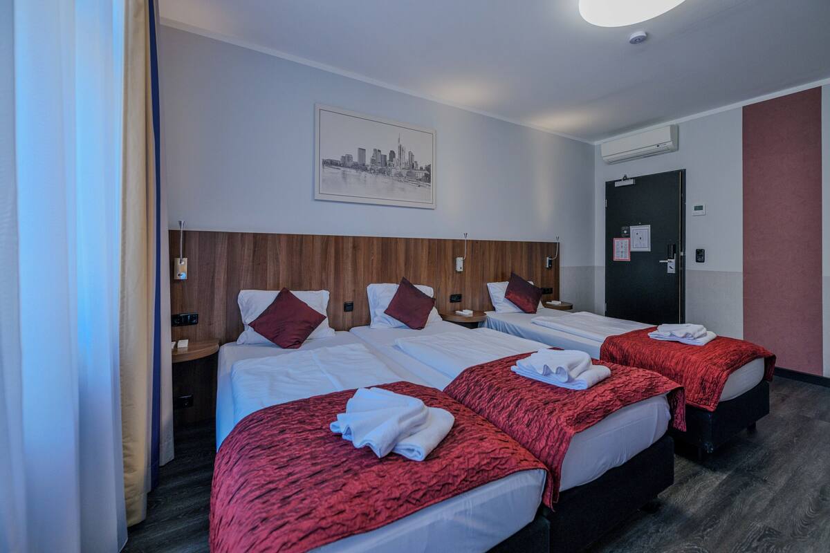 A hotel room with three beds, a cityscape artwork on the wall, a door with a security camera, and a nightstand with a phone.