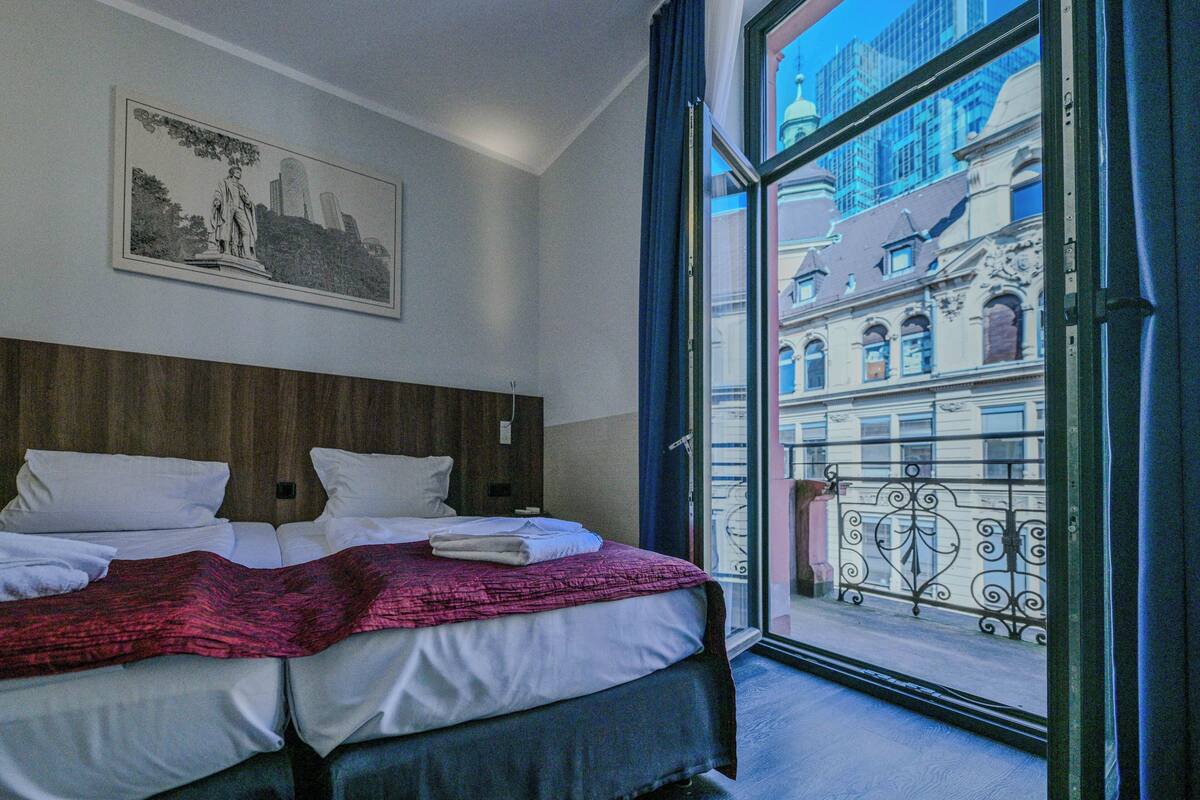 A hotel room with a bed, a framed picture on the wall, and a view of buildings through a window.
