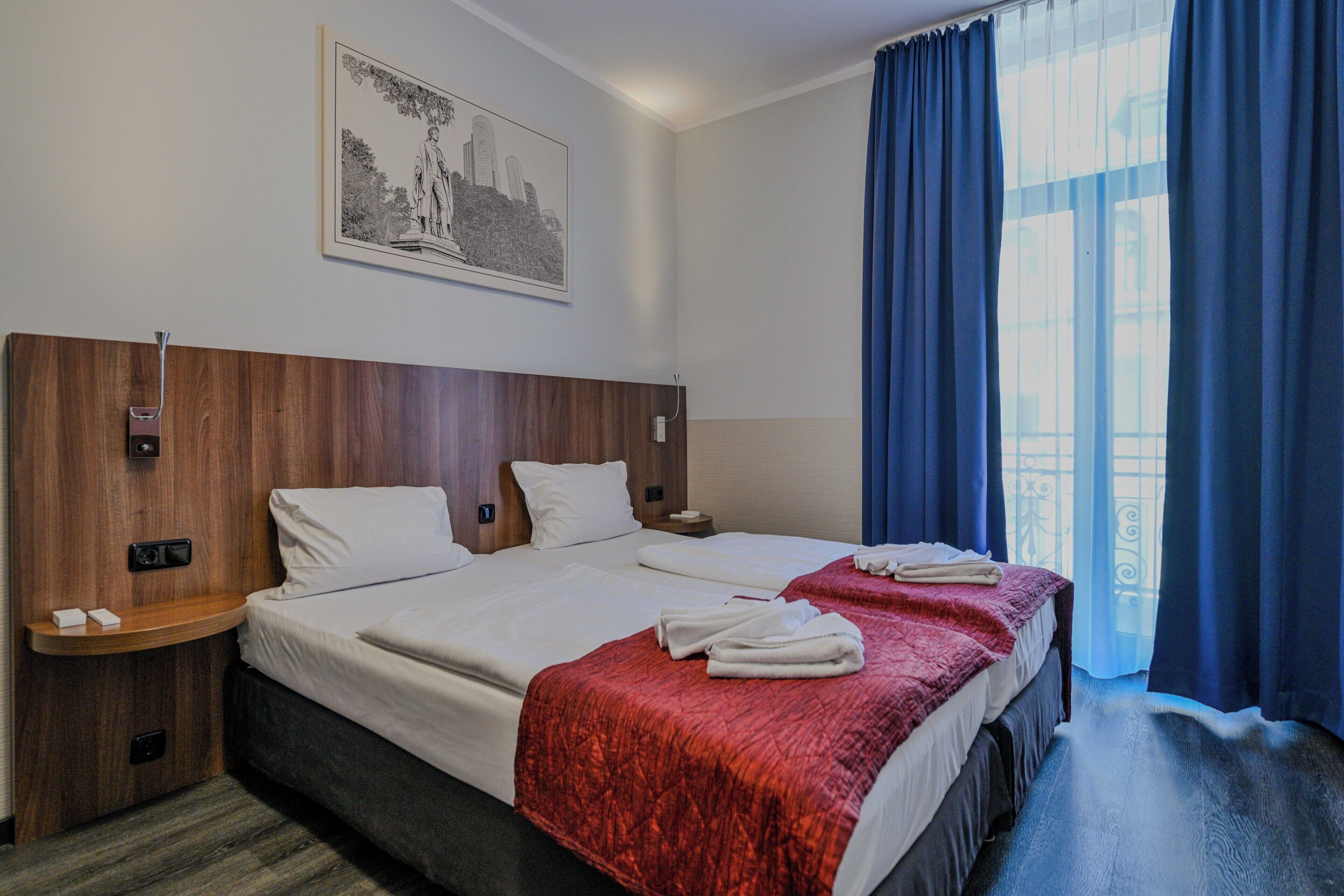 A hotel room with two beds, a wooden headboard, a nightstand, a wall-mounted light, and a window with blue curtains.