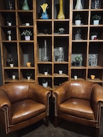 Una habitación con dos sillones de cuero marrón enfrentados, una estantería de madera con varios objetos decorativos y piso alfombrado.