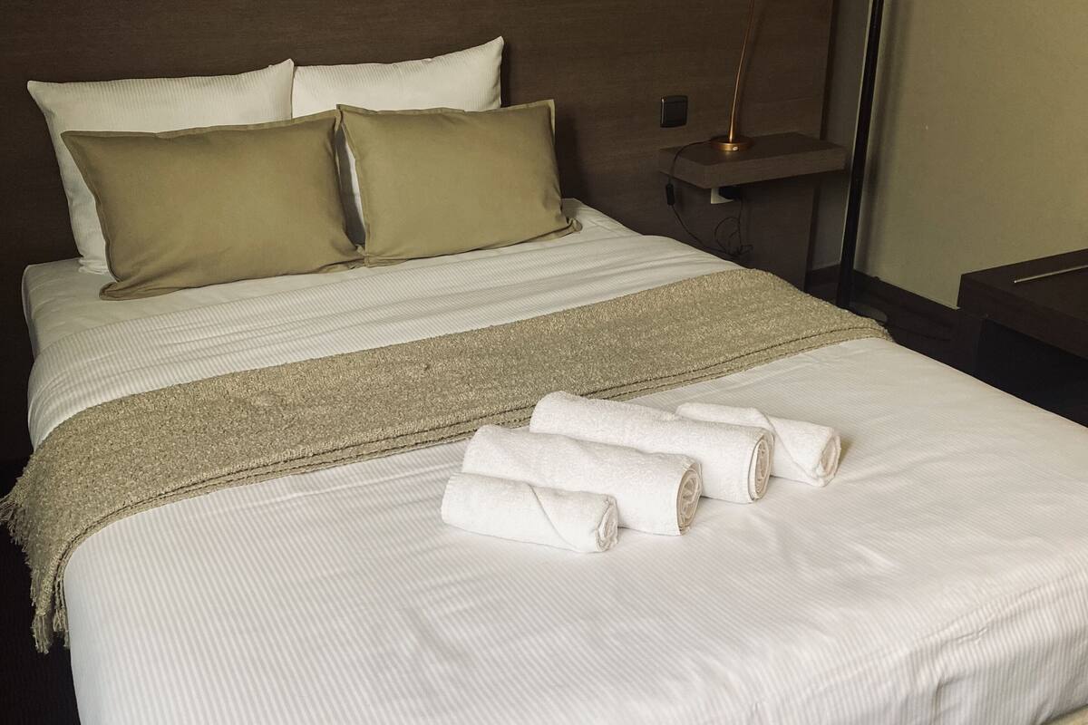 Una cama bien hecha con sábanas blancas, una manta color beige y toallas enrolladas.