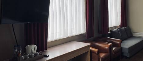 A hotel room with a flat-screen TV, a wooden desk, a leather sofa, and a grey couch.