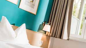 A hotel room with a teal wall, a bed with white linens, a bedside table with a lamp, and a window with curtains. - Hotel Heide Park (Ora)
