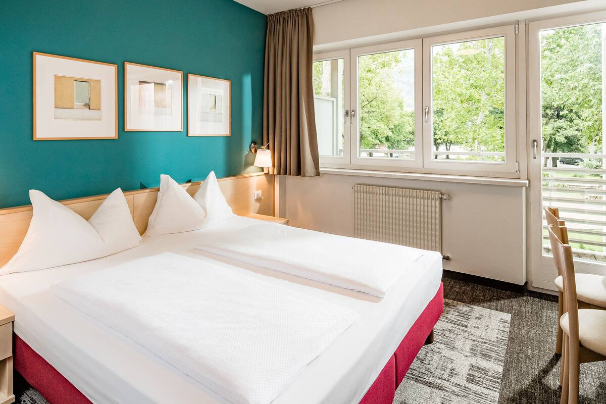 A hotel room with a large bed, a desk, a chair, a window with curtains, and framed pictures on the wall.