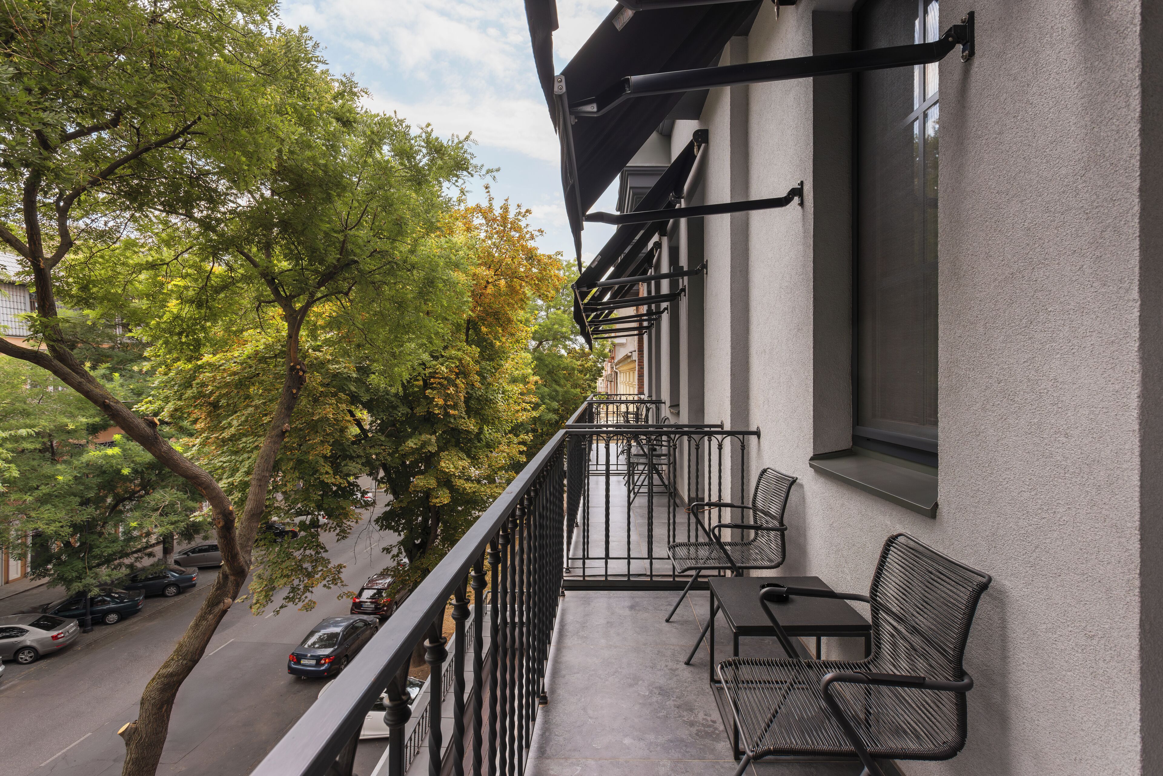 A balcony with outdoor seating, a view of a tree-lined street, and a building facade.