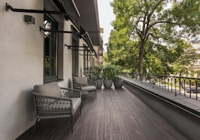 Terraza de madera con muebles de mimbre y plantas en macetas, bordeada por una barandilla y vista a la calle.