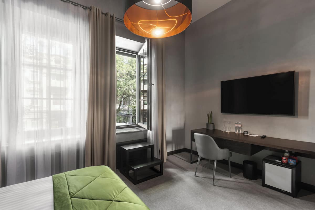 A modern hotel room with a bed, a desk, a chair, a TV, and a window with curtains.