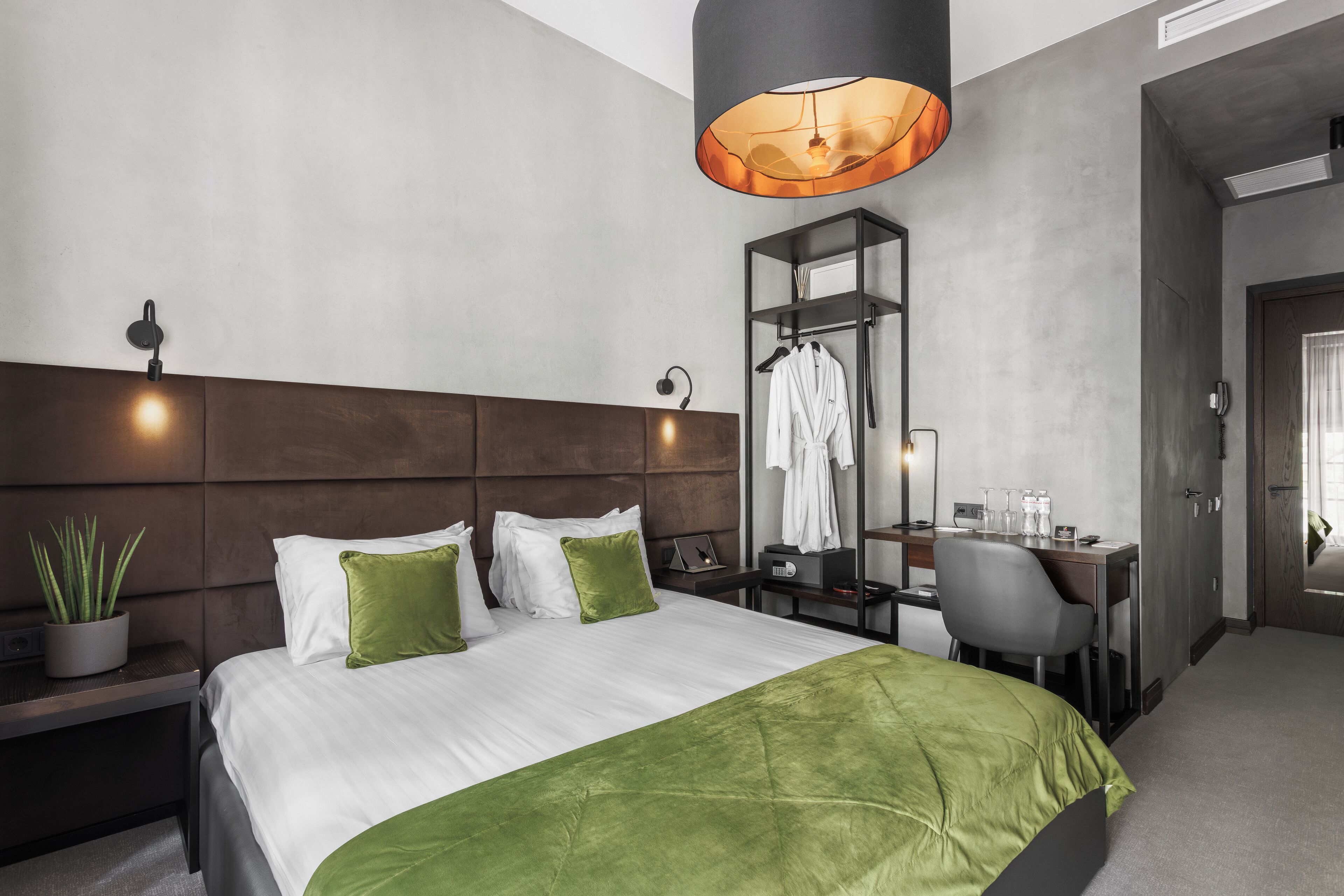 A modern hotel room with a large bed, a desk, and a chair. There is a potted plant on the left side of the bed, and a mirror on the wall above the desk.