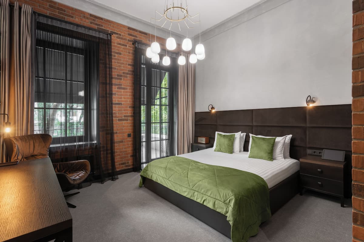 A modern bedroom with a brick wall, a large bed with green bedding, a wooden nightstand, and a hanging light fixture.