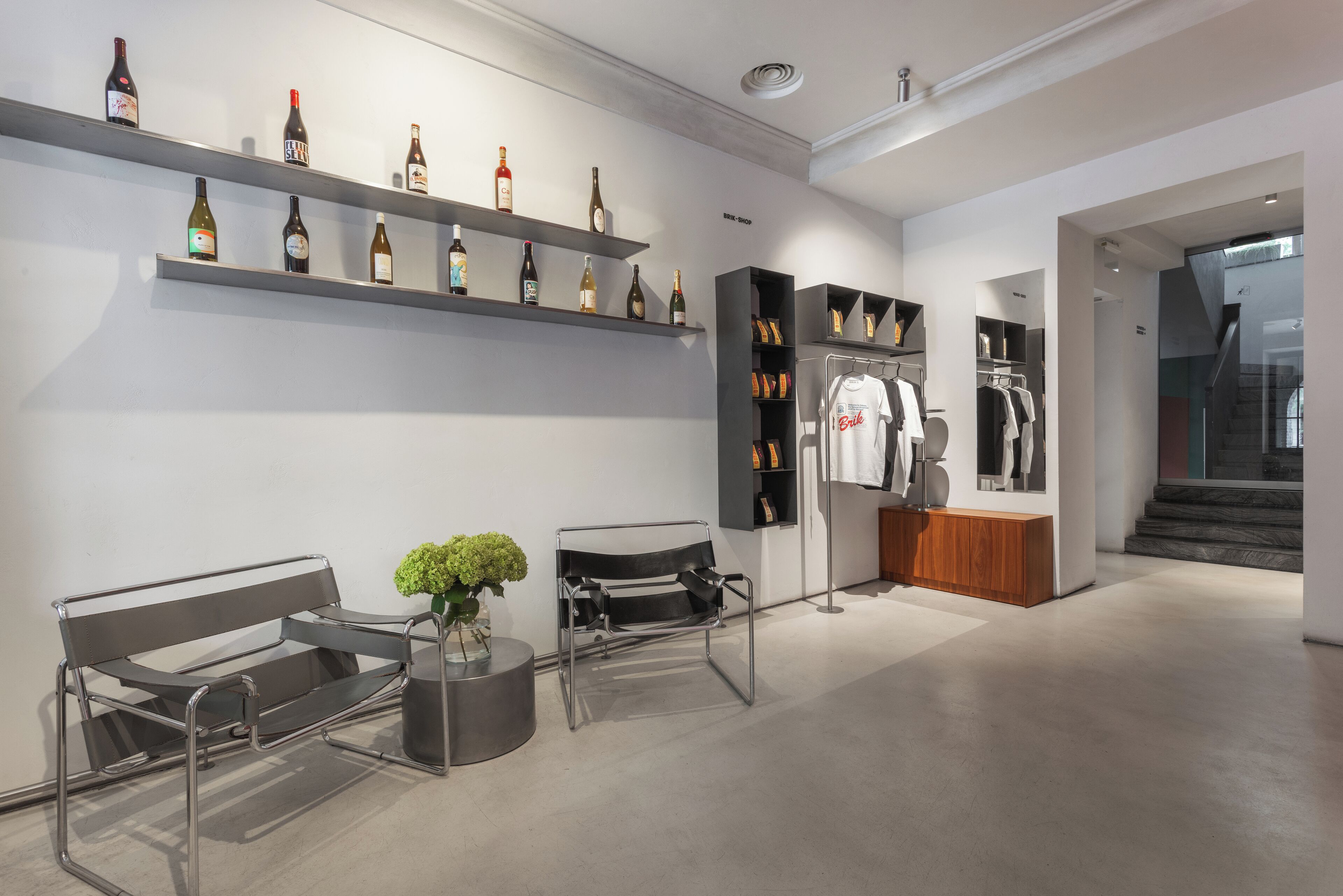 A modern interior with shelves of wine bottles, a bench, a small table with a vase, and a clothing rack.