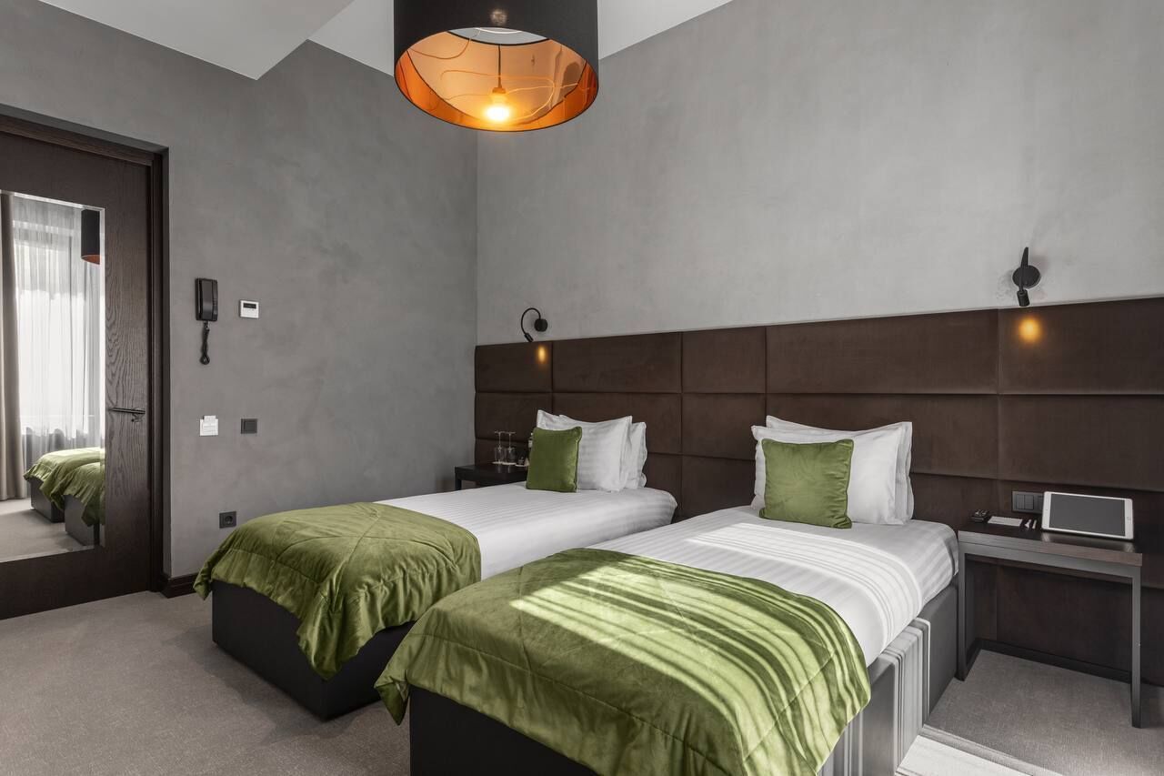 Una habitación de hotel moderna con dos camas, un espejo grande, una mesita de noche con lámpara y un aplique de techo.