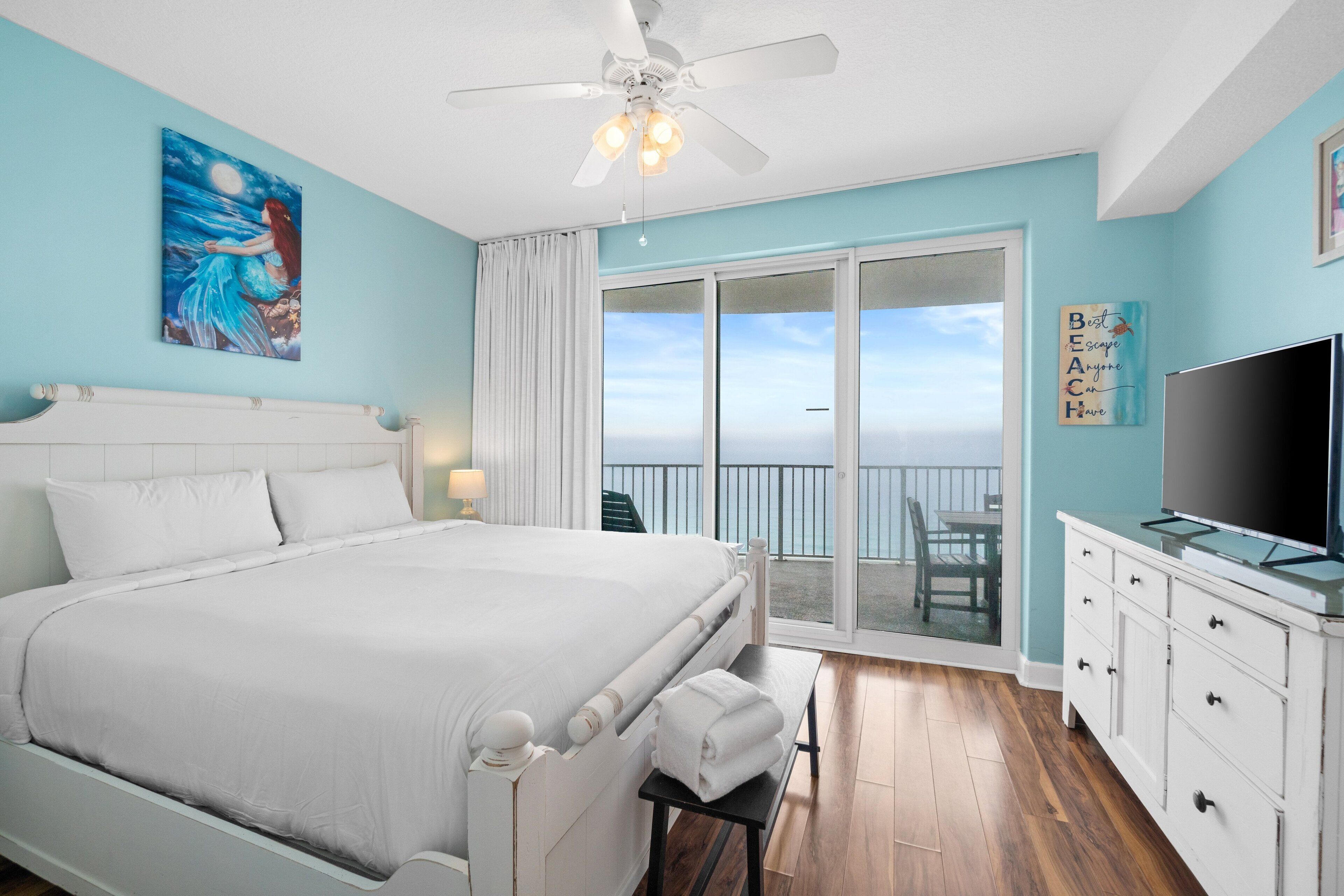 A bedroom with a bed, a ceiling fan, a TV, and a balcony with a view of the ocean.