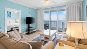 Ein Wohnzimmer mit Fernseher, Deckenventilator, großem Fenster mit Vorhängen und Blick aufs Meer.