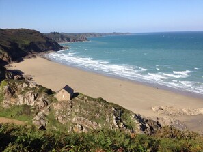 A coastal landscape with a sandy beach, rocky cliffs, and a small stone building.