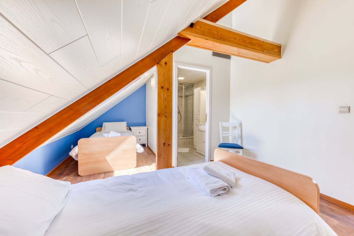 A bedroom with a sloped ceiling, a bed with white linens, a wooden headboard, a wooden bench, and a blue wall.