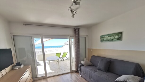 Ett rum med en grå soffa, en tv-apparat på ett träskåp, ett bord med stolar och utsikt över stranden genom en glasdörr.