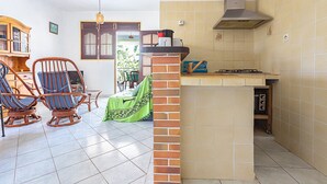 Una cocina con estufa a gas, un pilar de ladrillo y un comedor con sillas de mimbre y mantel verde.