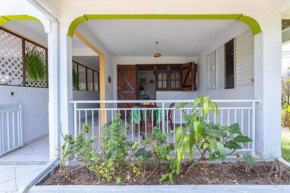 A covered porch with a white railing, a small garden area with plants, and a view into a house with a dining area and kitchen visible. - Le Verger de Charles - jardin arboré - Petit Canal Guadeloupe (Petit-Canal)
