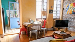Un coin repas avec une table dressée pour quatre personnes, des chaises rouges et blanches, un téléviseur et un tableau accroché au mur.