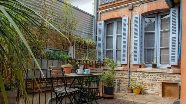 Une terrasse en bois avec une table et des chaises, entourée de plantes en pot et d’un mur de briques.