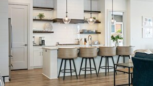 Cocina moderna con armarios blancos, una isla central con taburetes de bar, iluminación colgante y vistas al área de estar contigua.