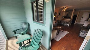 A porch with three Adirondack chairs, a wooden table, and a window with blinds. - Newly Renovated Hilton Head Resort Beach Condo - 2 Bedroom - Private Balcony (Hilton Head Island)