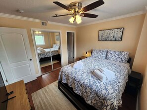 Un dormitorio con cama, ventilador de techo, mesita de noche, espejo y una puerta.