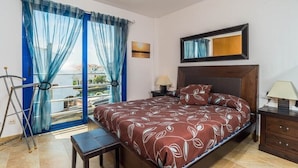 Une chambre à coucher avec un lit, une table de chevet, une lampe, une fenêtre avec des rideaux et une vue sur l’extérieur.