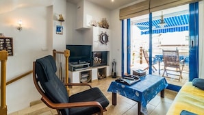 Un salon avec un téléviseur, une chaise en bois, une table recouverte d’une nappe bleue et un balcon agrémenté de rideaux bleus et de mobilier d’extérieur.