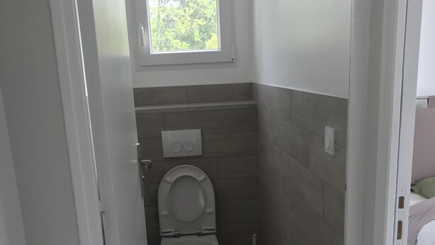 Une petite salle de bain étroite, avec des toilettes, une fenêtre et une brosse à récurer les toilettes.