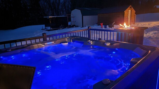 A hot tub with blue lighting, surrounded by a metal fence, in a snowy outdoor setting with a wooden structure in the background. - Piece of Paradise / Private Hot Tub (Holcombe)