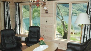 A cozy room with a wooden interior, a leather sofa, two chairs, a small table with a vase of flowers, and large windows offering a view of a lake and forest. - Holiday Home in Olden (Stryn)