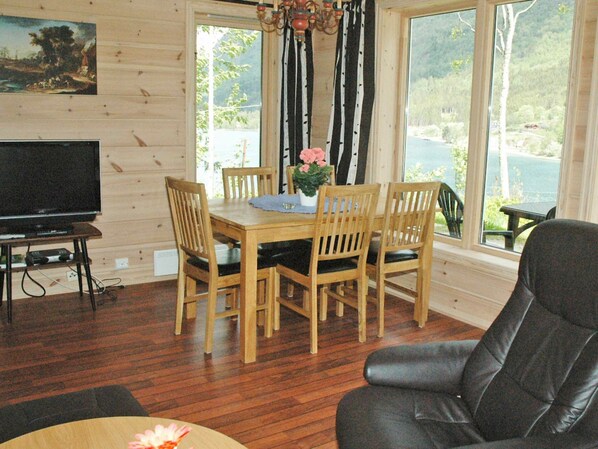 A wooden interior with a dining table, chairs, a television, and a view of a lake through large windows.