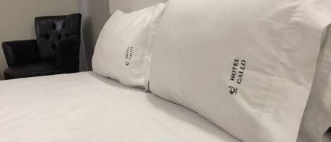 A bed with white bedding and pillows featuring the Hotel Gallo logo.