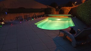 A swimming pool with a curved edge, surrounded by a tiled patio area with chairs and a hose.