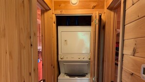 Un pasillo estrecho con paredes de madera y un aplique en el techo. En el centro se ve un electrodoméstico blanco, posiblemente una lavadora y secadora.