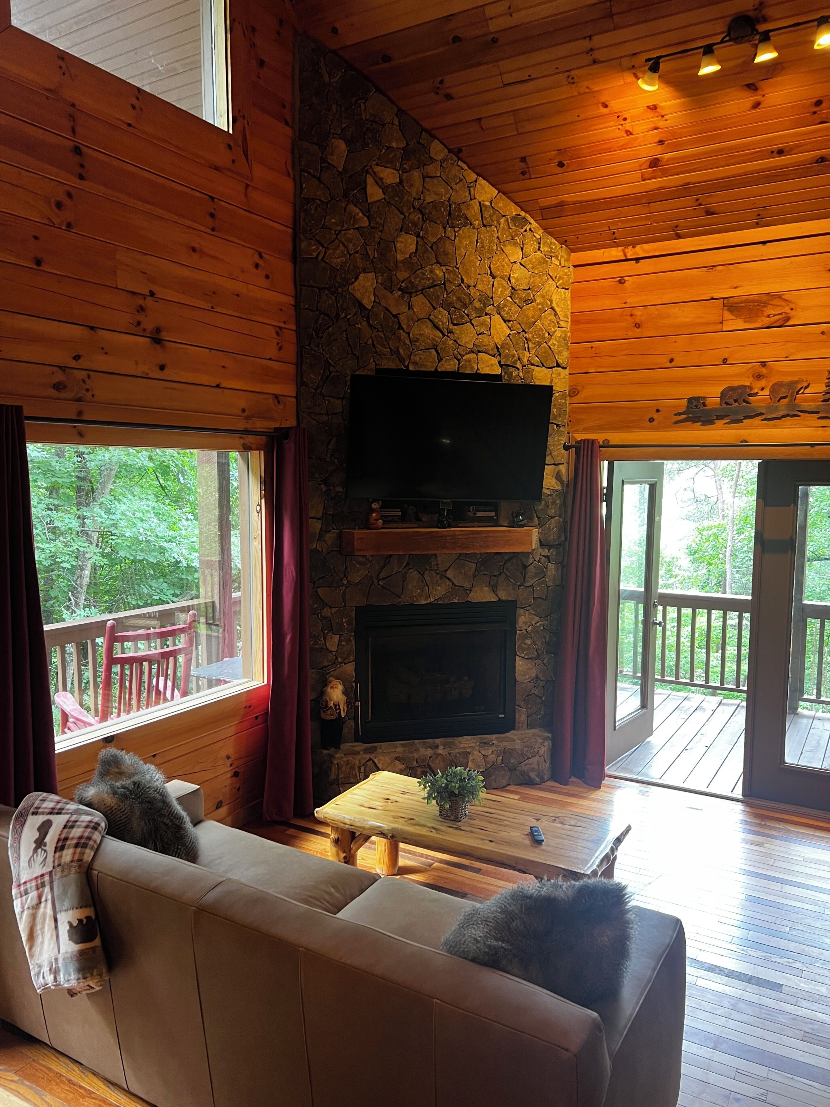 A cozy cabin interior with a stone fireplace, a flat-screen TV mounted above it, a wooden ceiling, and a large window overlooking a deck.