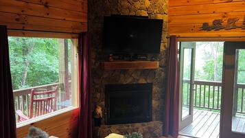 Interior acogedor de una cabaña con chimenea de piedra, televisor de pantalla plana montado encima y techo de madera, con un gran ventanal con vistas a una terraza.