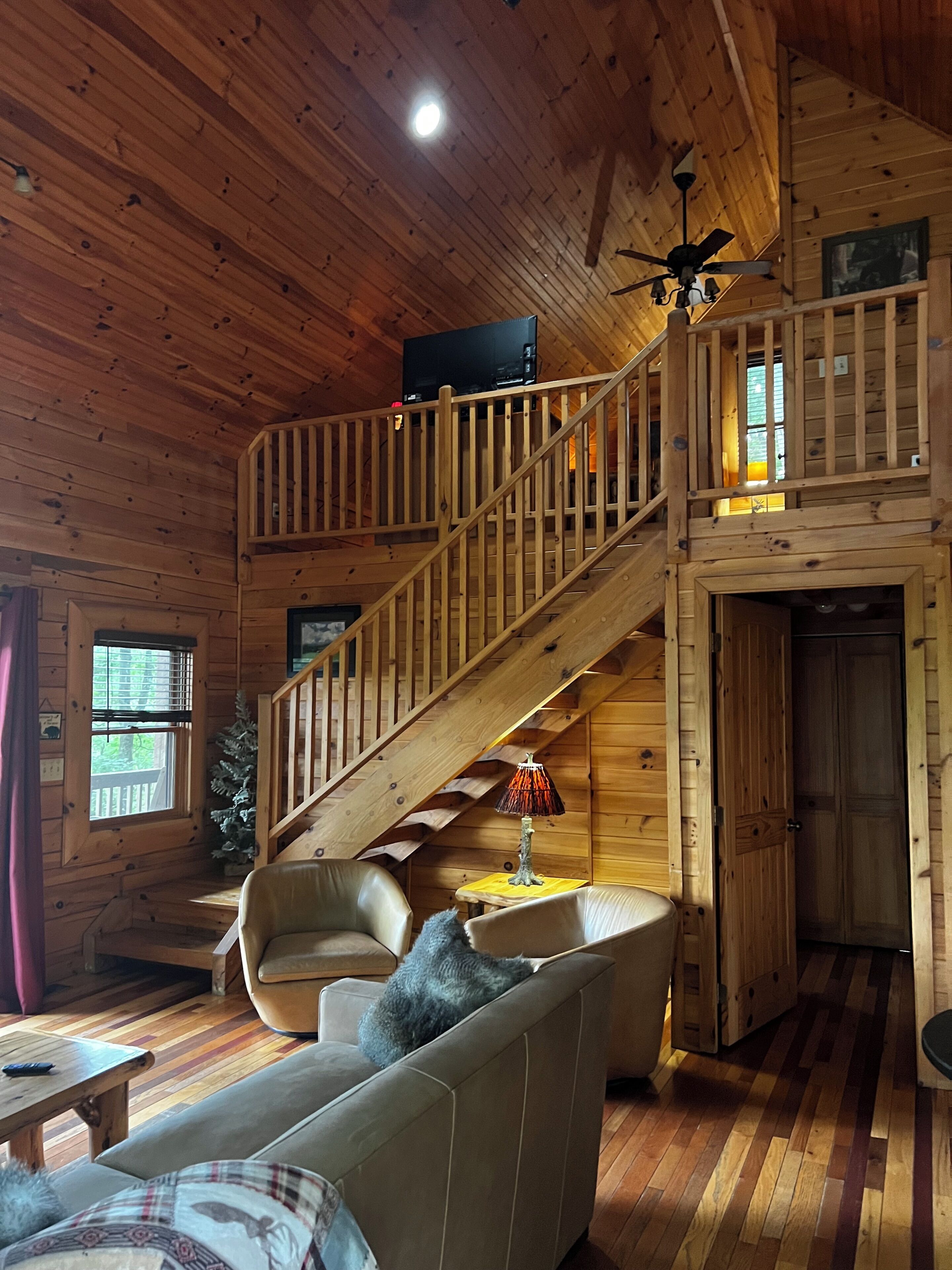 Interior de una cabaña de madera con una escalera, un sofá, una lámpara y un televisor en el nivel superior.
