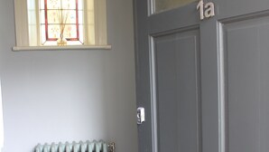 Una puerta gris oscuro cerrada con una placa de número '1a', un radiador y una ventana con un diseño de vitral.