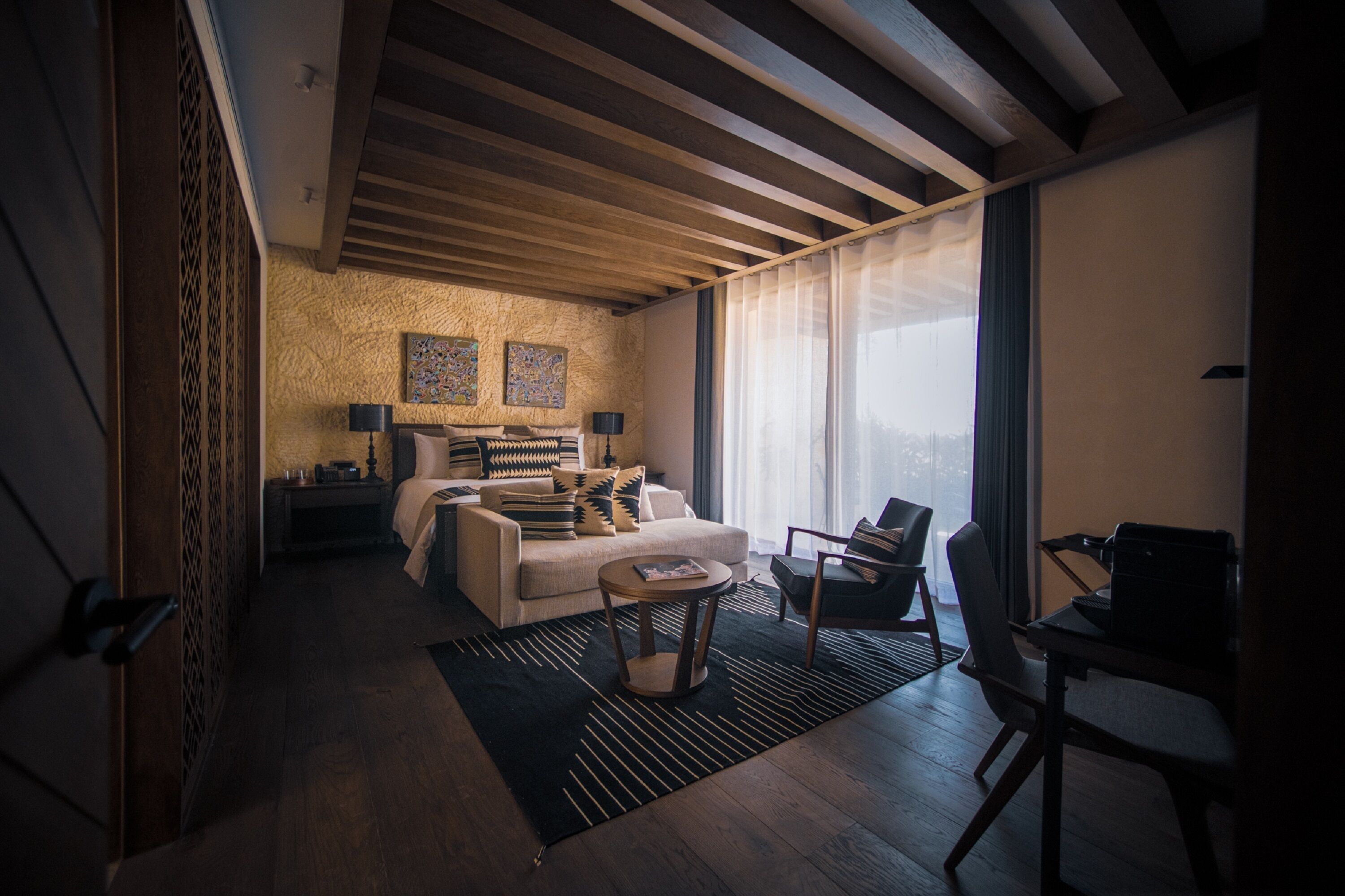 A modern living room with a sofa, coffee table, and chairs, wooden beams on the ceiling, and large windows with sheer curtains.