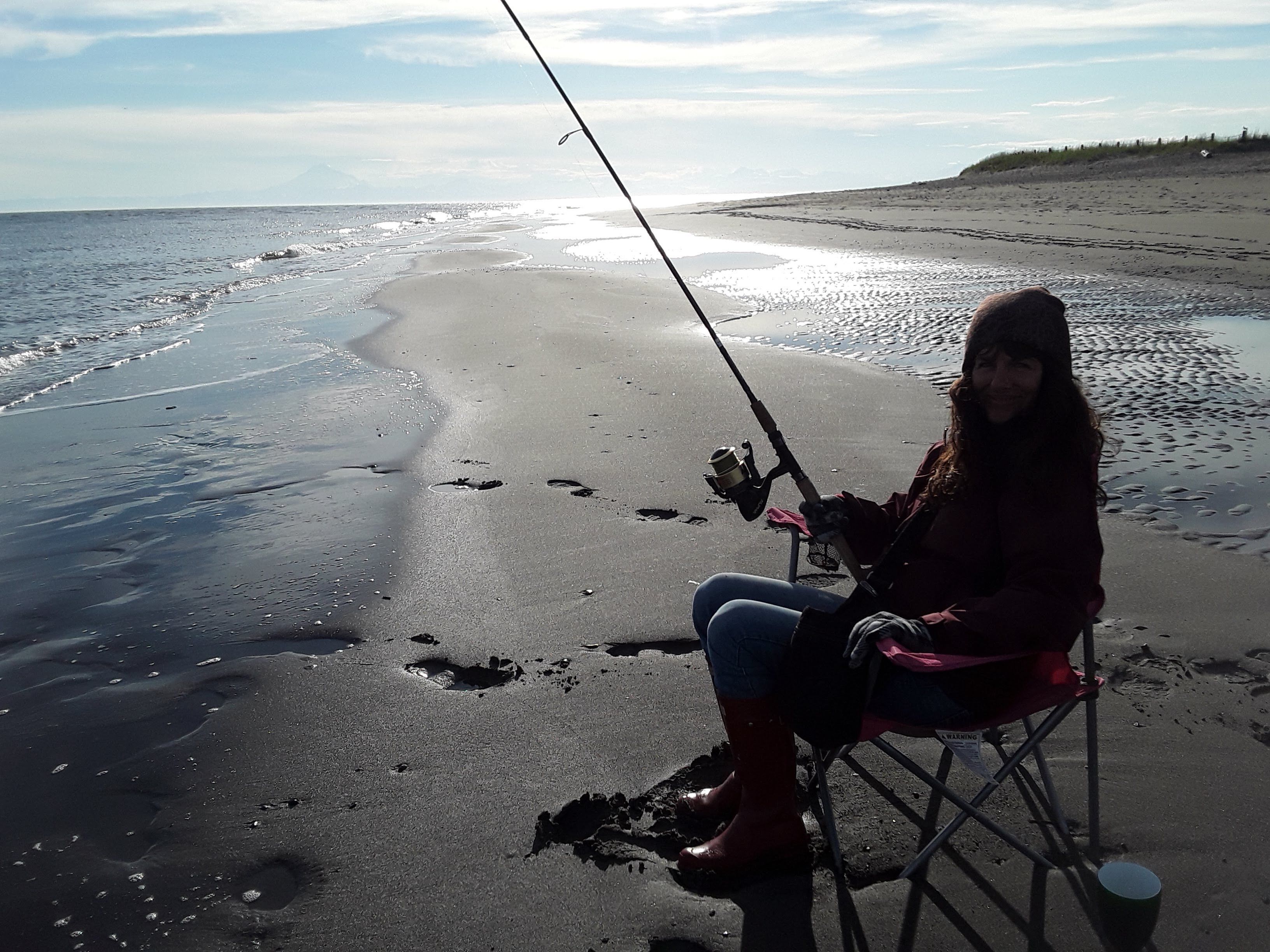 Una persona sentada en una silla de playa sosteniendo una caña de pescar, con un mar tranquilo y un cielo despejado al fondo.