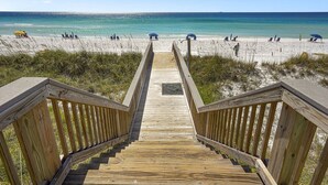 A wooden boardwalk leading to a beach with clear blue water and people relaxing under umbrellas.
