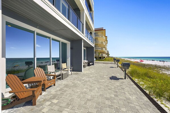 Une terrasse donnant sur la plage, avec des chaises et des tables en bois, de grandes fenêtres offrant une vue sur la plage et l’océan, et un bâtiment avec des balcons en arrière-plan.