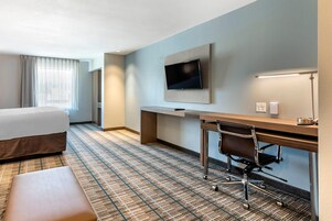 A hotel room with a bed, a desk with a chair, a flat-screen TV mounted on the wall, and a lamp on the desk.