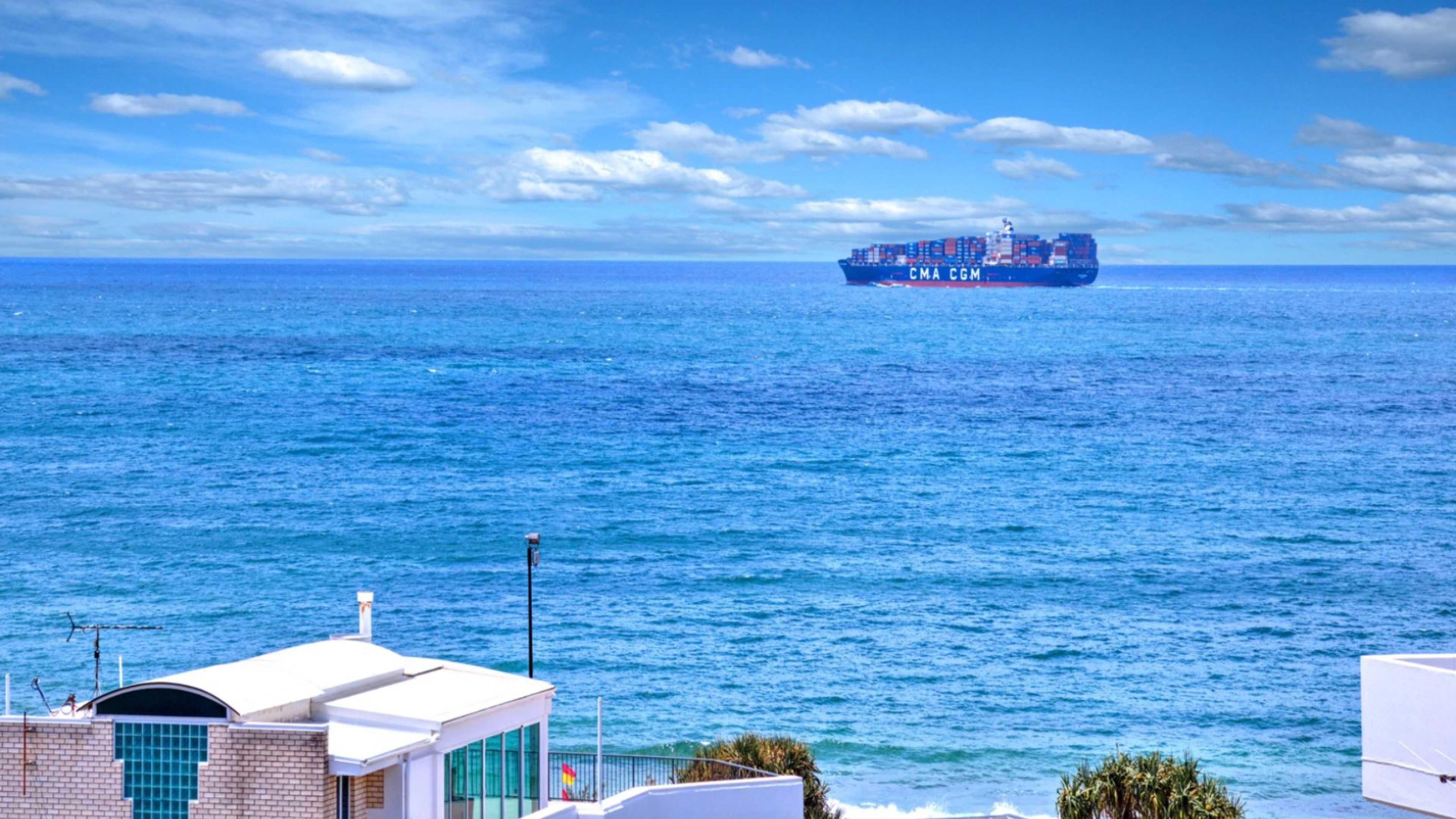 A coastal view with a large cargo ship in the distance, a building with a white facade, and a clear blue sky.