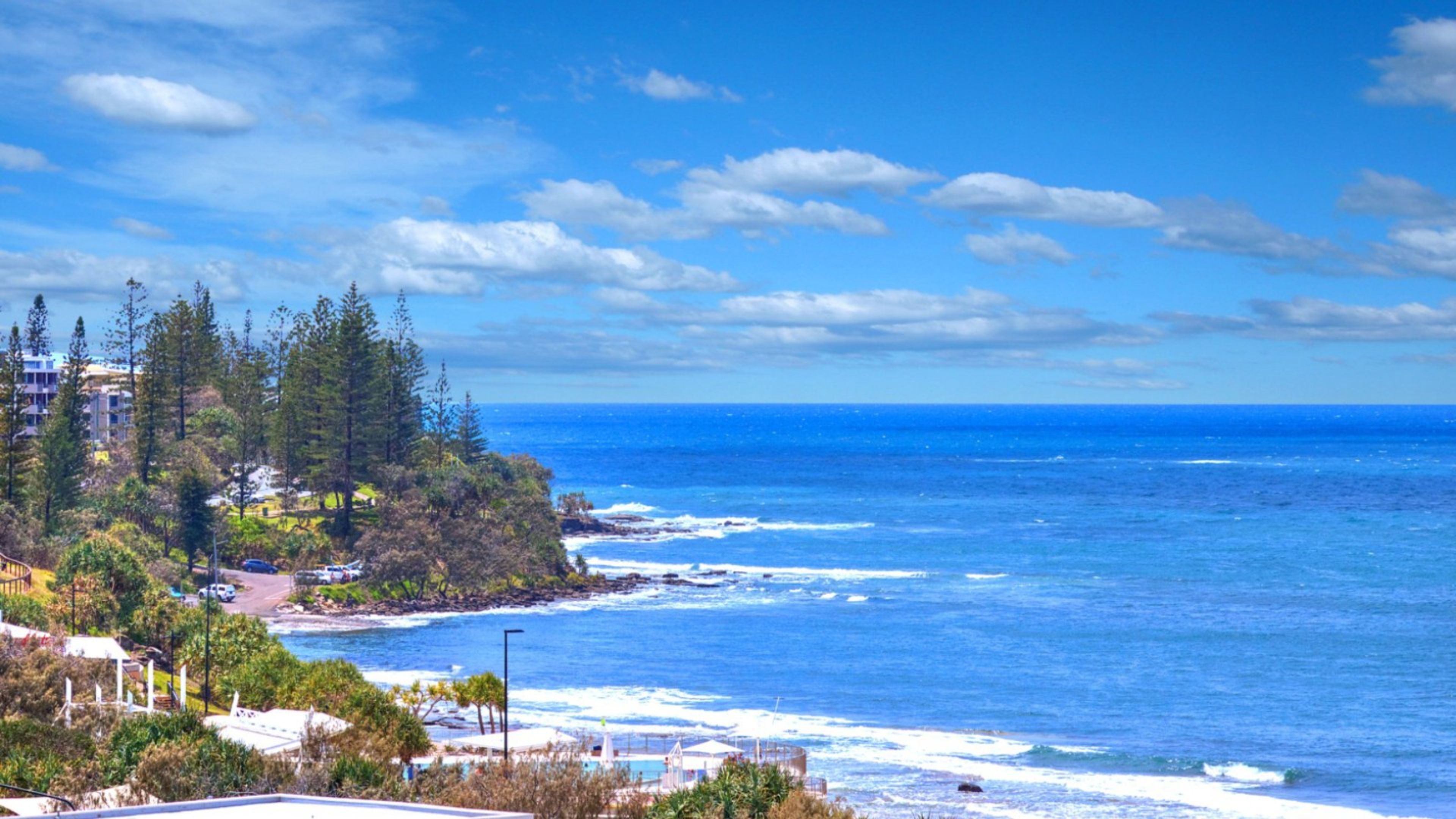 A coastal view with ocean waves, a clear sky, and a residential area with buildings and trees.